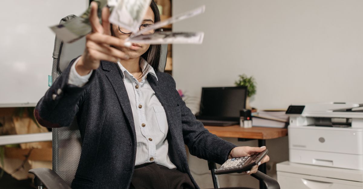 A businesswoman in a suit throws money while calculating finances in a modern office.