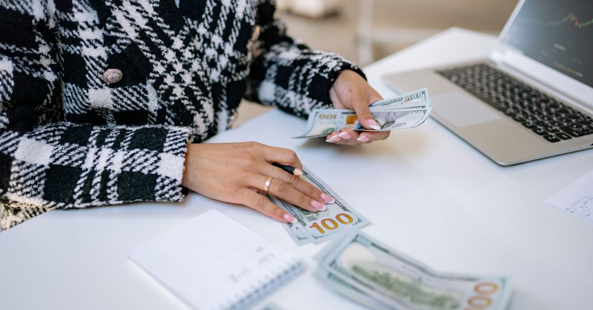 Close-up of a woman counting hundred-dollar bills at a desk with a laptop, focusing on finance and work.
