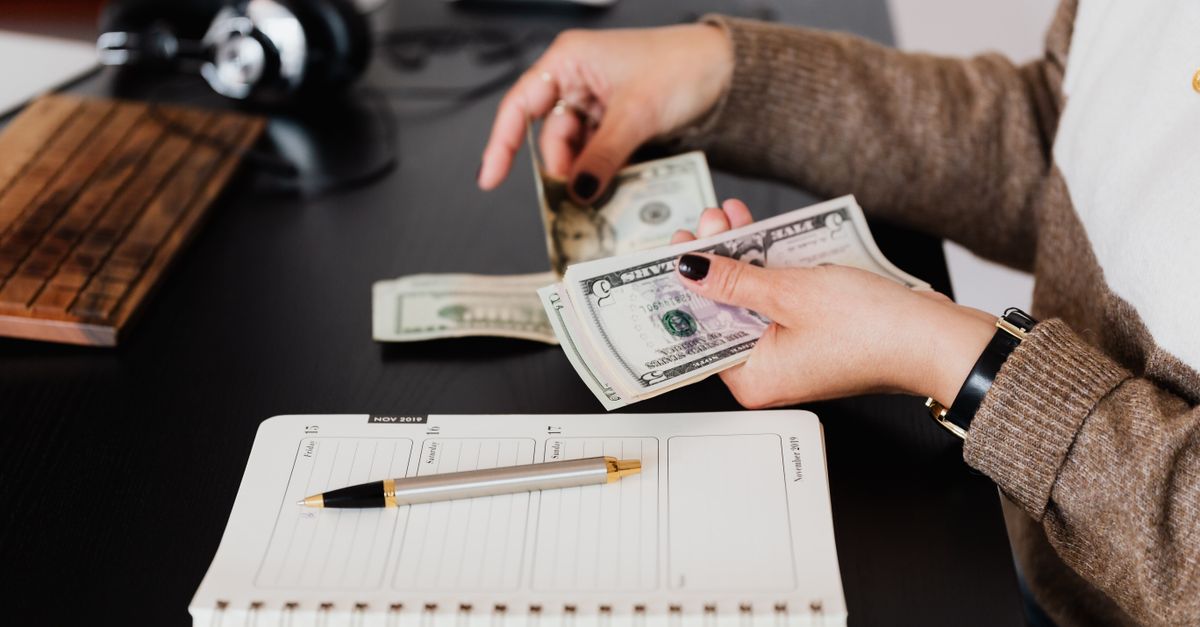 Side view of crop anonymous smartly dressed female entrepreneur counting dollar banknotes while sitting at modern office desk with planner and pen