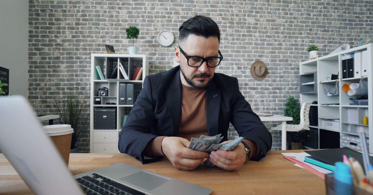 Businessman counting cash at a desk in a modern office setting with a laptop and documents.
