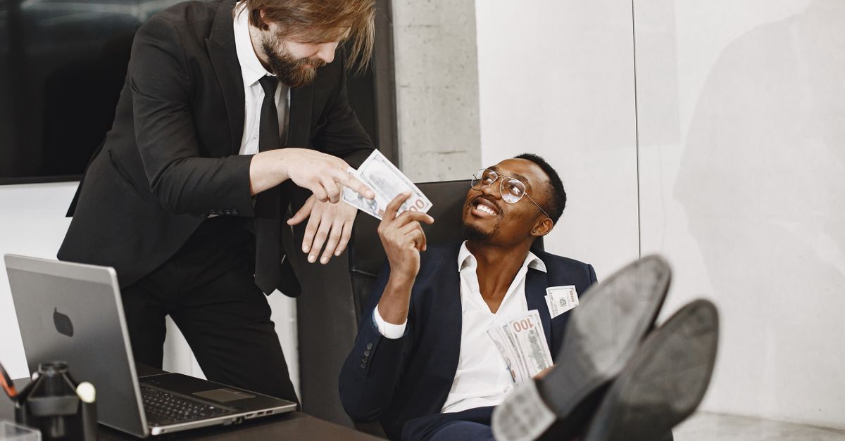 Two businessmen in suits exchanging money with smiles in a modern office environment.