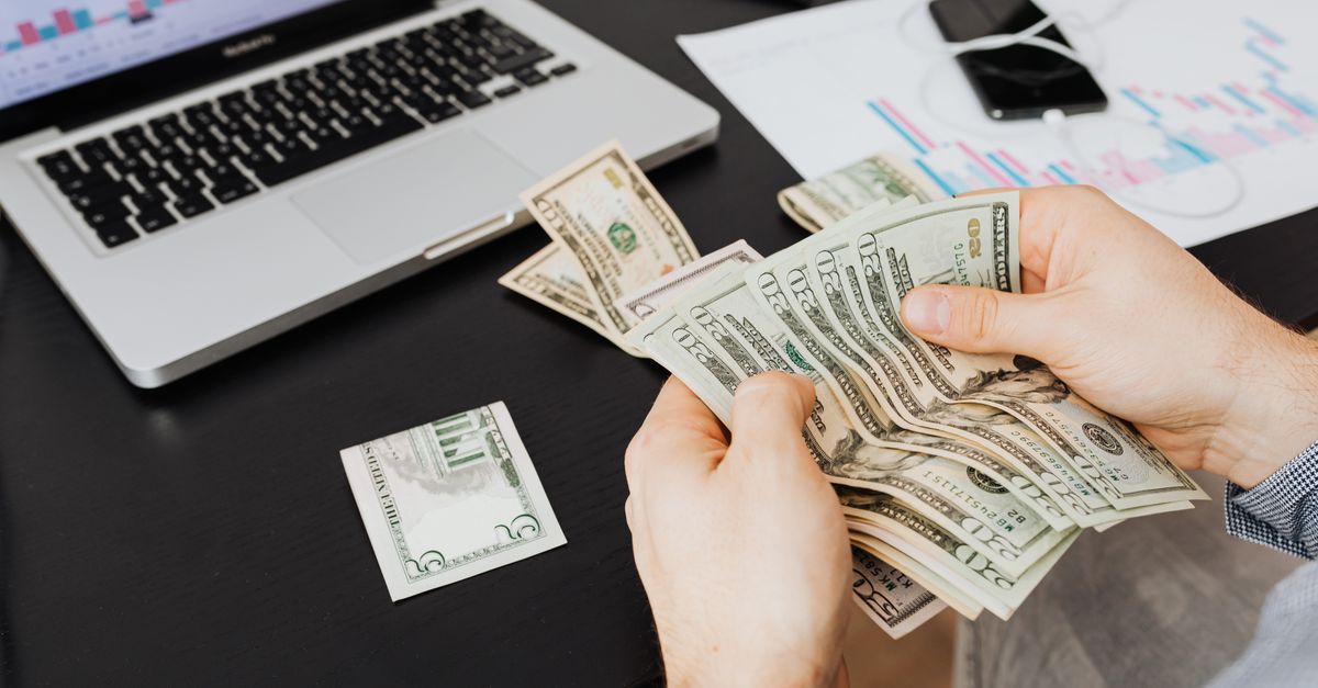 Person counting dollar bills at desk with laptop and financial charts, symbolizing finance management.