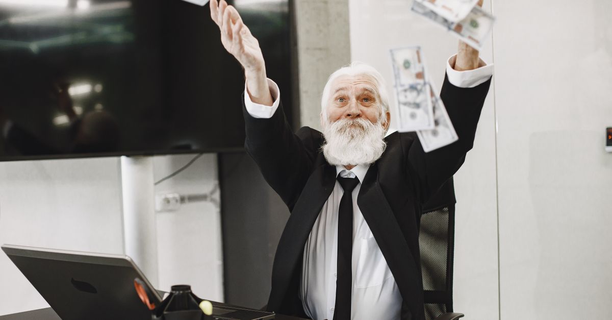 A joyful senior businessman in a suit throws money in an office setting, expressing financial triumph.