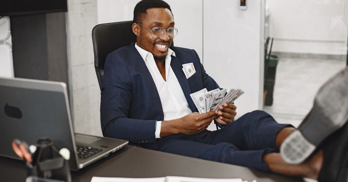 Businessman in a suit counts cash with a smile, relaxing in a modern office setting.